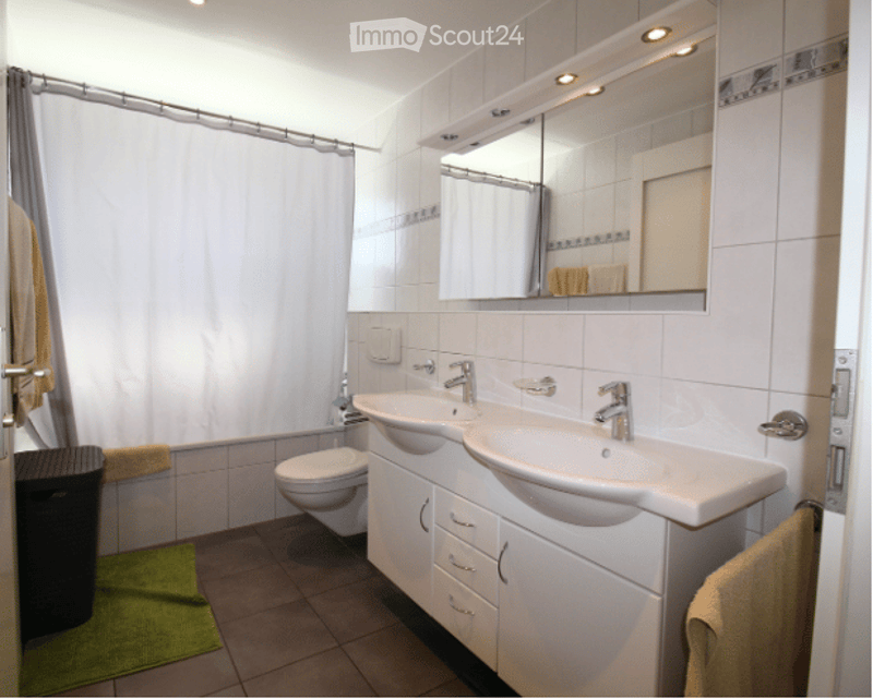 Modern bathroom with double sink vanity, large mirror, toilet, and shower area with a white curtain. White tiles and a green bath mat are visible.