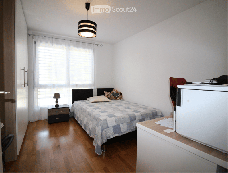 A small, tidy bedroom with a single bed, nightstand, desk, and a small refrigerator. The room features wooden flooring, white walls, and a window with white curtains.