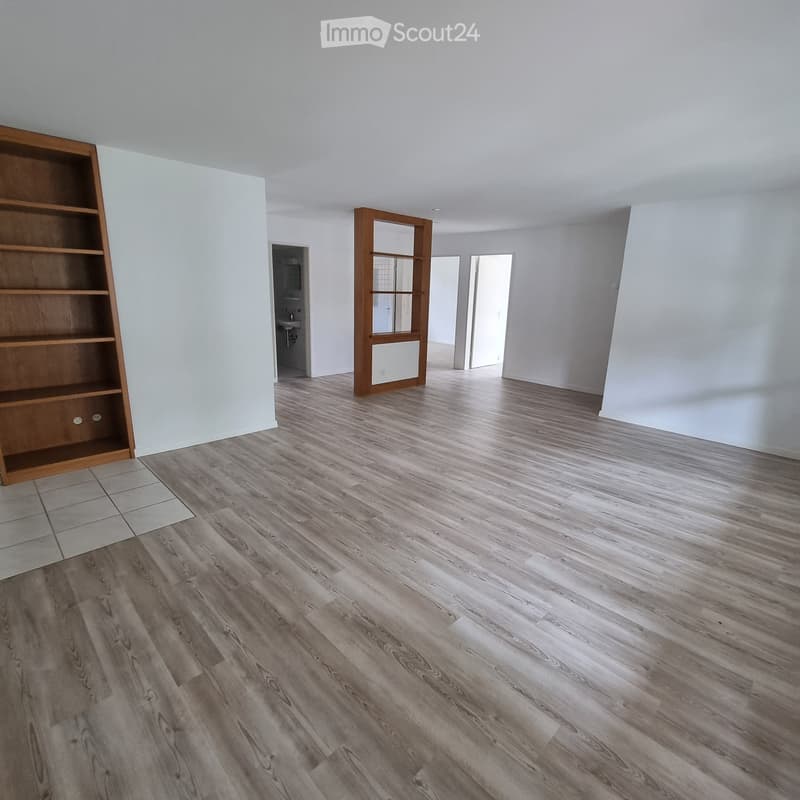 Empty room with light-colored walls, wooden flooring, and a wooden shelving unit on the left side. A doorway leads to another room with a visible bathroom.