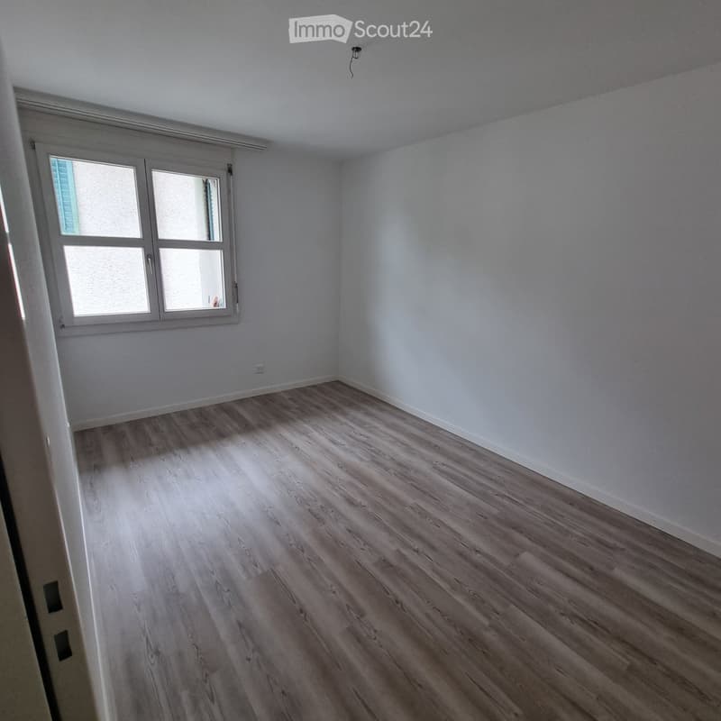Empty room with light wooden flooring and white walls, featuring a window with white frame and a door leading to another space.