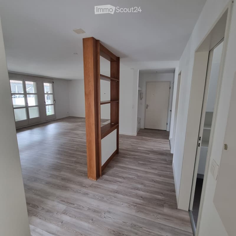 Empty hallway with light wooden flooring, white walls, and a wooden shelving unit on the left side. Natural light enters through a large window at the far end.