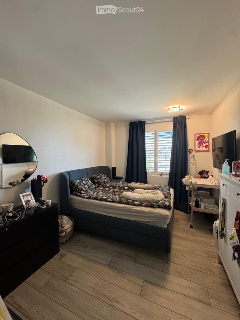 Cozy bedroom with wooden flooring, a large bed with floral bedding, a nightstand with a lamp, a mirror, and a small table with a chair near the window.
