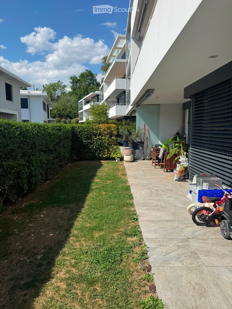 A well-maintained garden area with a concrete pathway leading to a patio, featuring a small lawn, potted plants, and a modern building in the background.