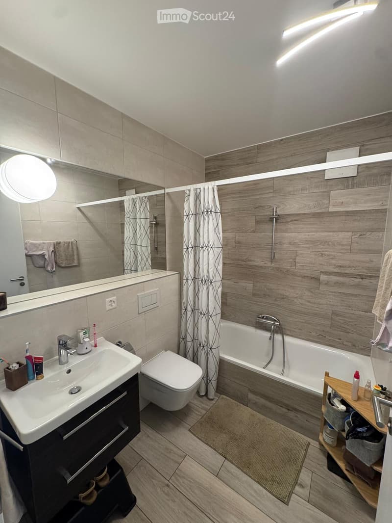 Modern bathroom with a sink, toilet, bathtub, and large mirror. Features wooden wall paneling and a shower curtain.