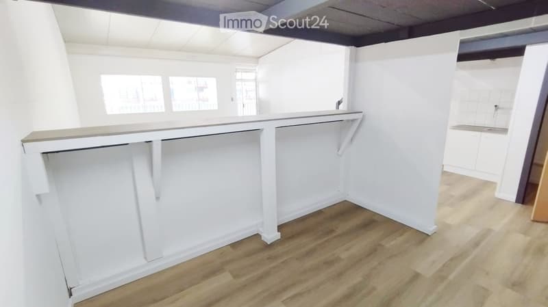 Empty room with white walls, wooden flooring, and a large white counter with built-in storage. Windows provide natural light.
