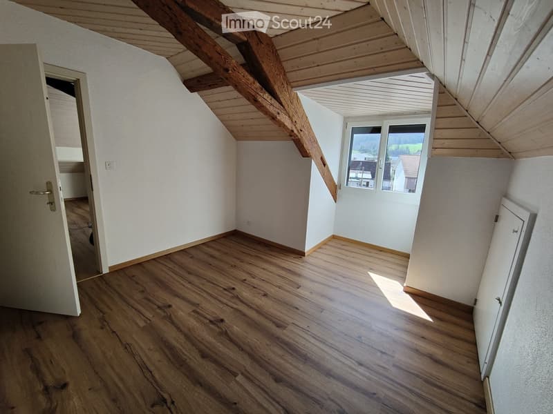 Empty room with wooden flooring, white walls, and exposed wooden beams on the ceiling. Features a large window with a view of the outside and a door leading to another room.