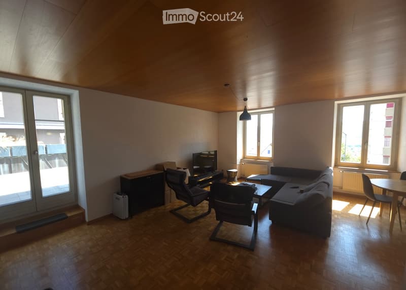 Modern living room with wooden flooring, large windows, and a balcony door. Features a sectional sofa, coffee table, chairs, and a television.