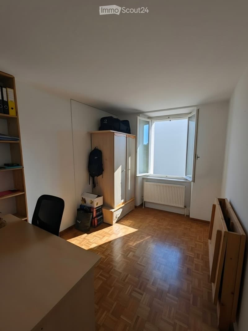 A small, furnished room with wooden flooring, a chair, a cabinet, a shelf, a radiator, and some personal items like a backpack and books.