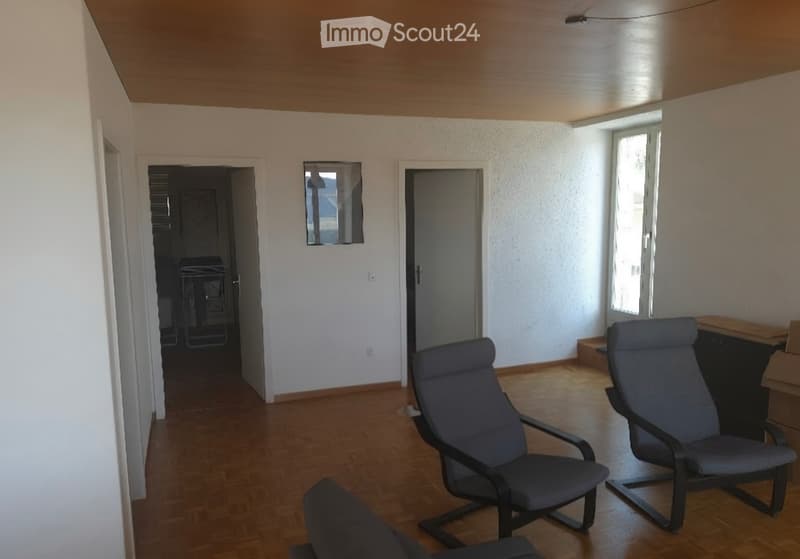 A small, unfurnished room with wooden flooring, white walls, and two chairs near a window. A small table is visible in the corner, and a mirror is mounted on the wall.