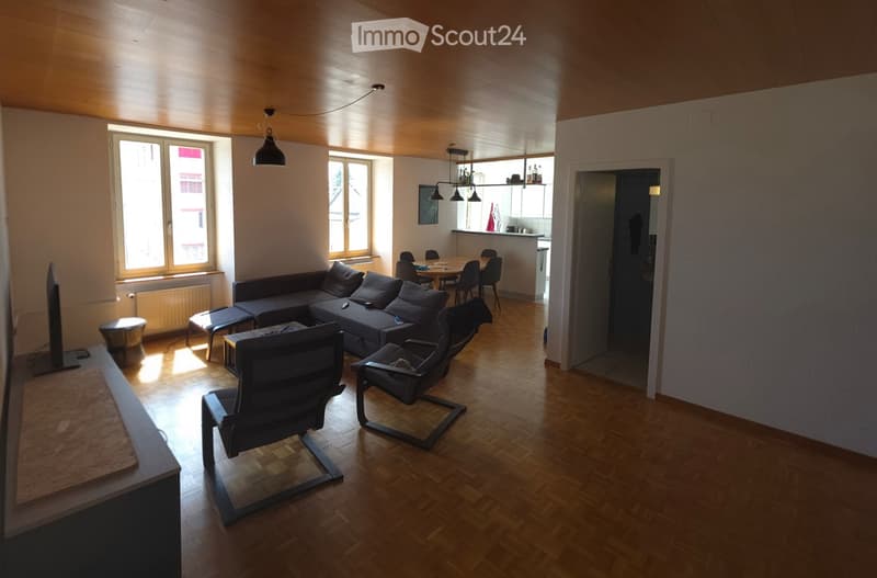 Bright living room with wooden flooring, large windows, a sectional sofa, chairs, a coffee table, and a TV stand.
