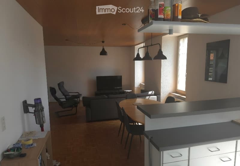 A modern living room with a sofa, a dining table with chairs, a television, and a hanging lamp. The room features wooden flooring and large windows allowing natural light to fill the space.