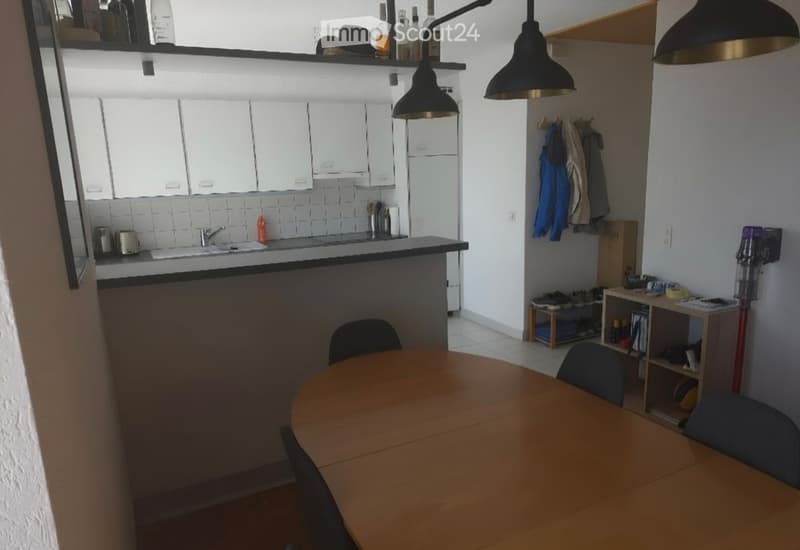 A modern kitchen and dining area with white cabinets, a wooden dining table, and a sink area with a faucet. Shelves hold various items, and a coat rack is visible.