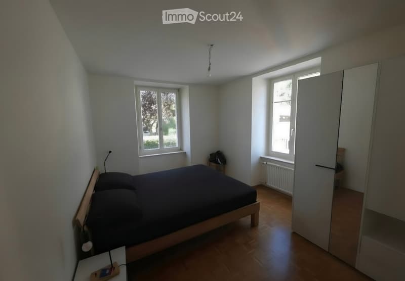 A tidy bedroom with a wooden bed, nightstand, and wardrobe. The room features wooden flooring, white walls, and large windows letting in natural light.
