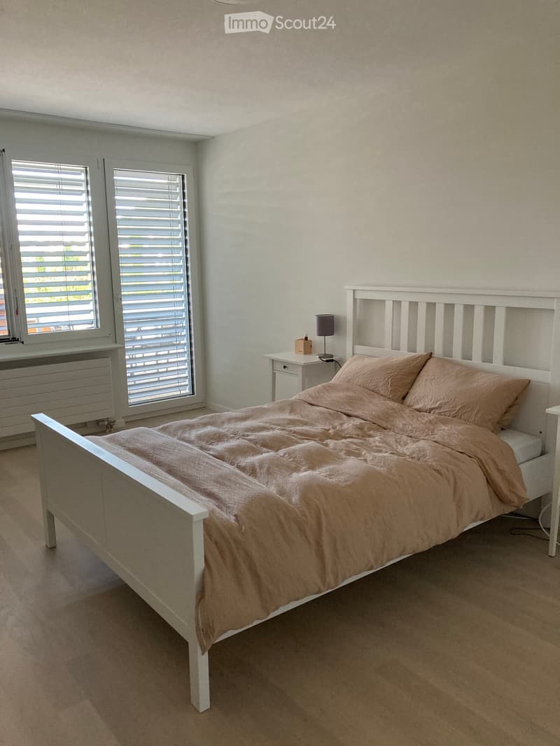Bedroom with a white wooden bed, beige bedding, a nightstand with a lamp, and two windows with blinds.