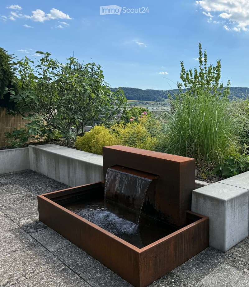 A modern water fountain with a sleek, dark metal design, surrounded by concrete planters and lush greenery, set against a scenic backdrop of hills and a clear blue sky.