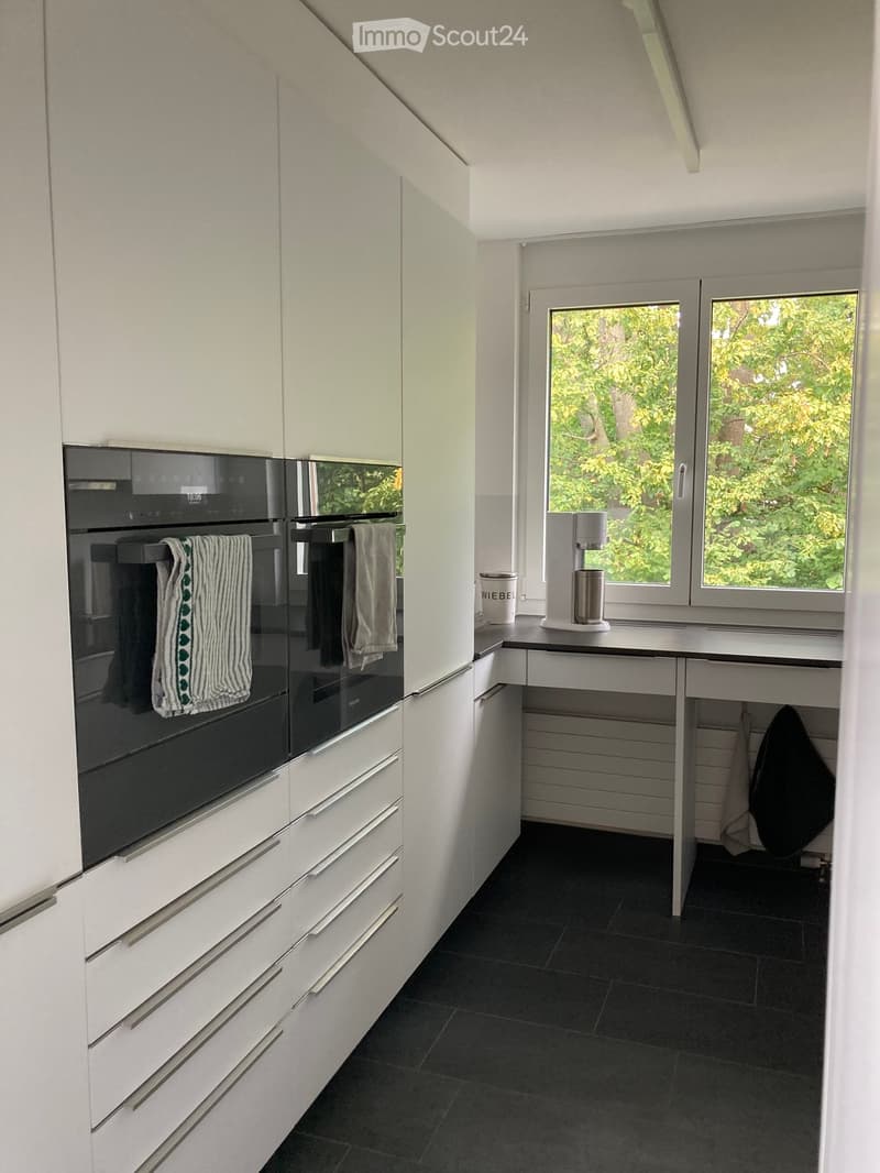 Modern kitchen with white cabinets, black oven, coffee maker on counter, and large windows showing greenery outside.