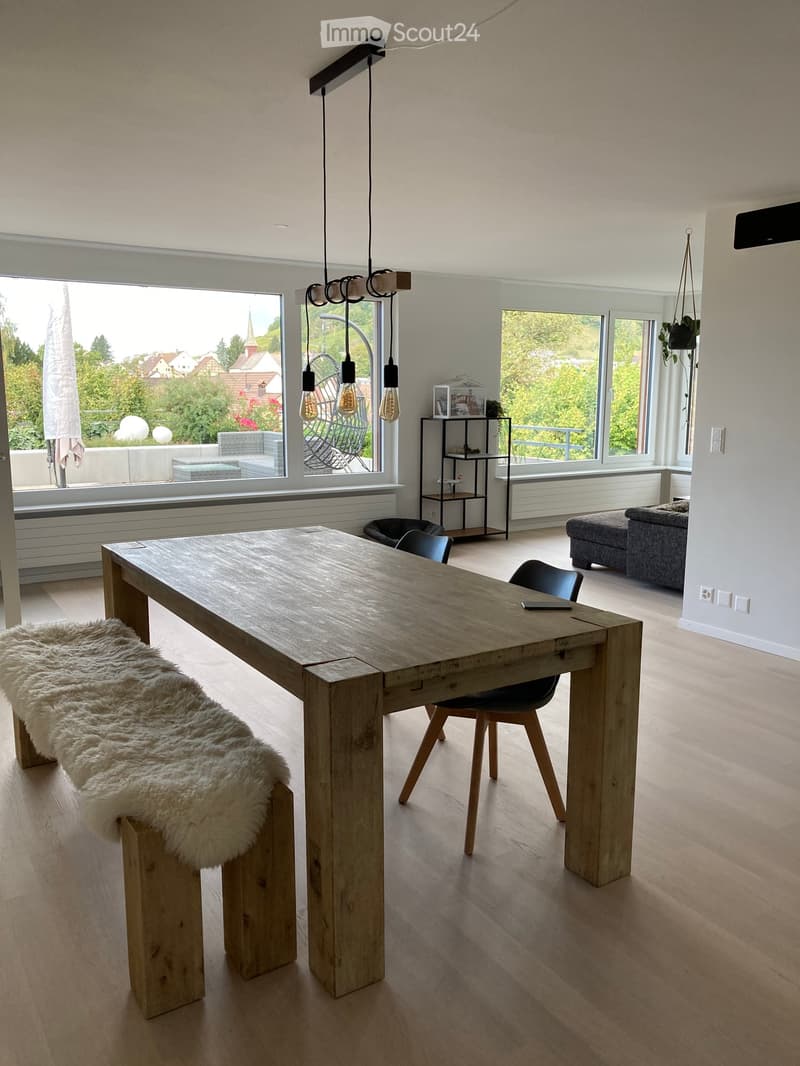 Bright, modern dining area with a large wooden table, bench, chairs, and a hanging lamp. Large windows offer a scenic view of the outdoors.