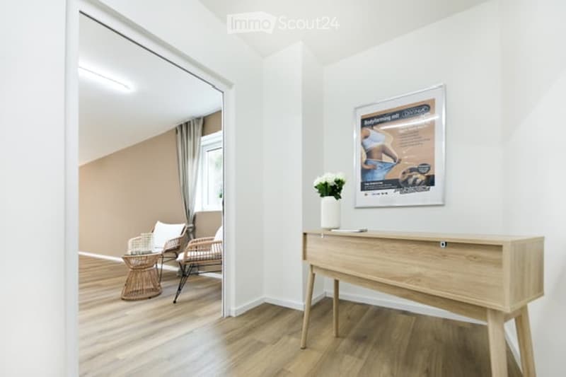 A modern, minimalist hallway with light wooden flooring, white walls, and a sleek console table. A framed poster is displayed on the wall, and a small vase with white flowers sits on the table. A wicker chair and small round table are visible in the adjac