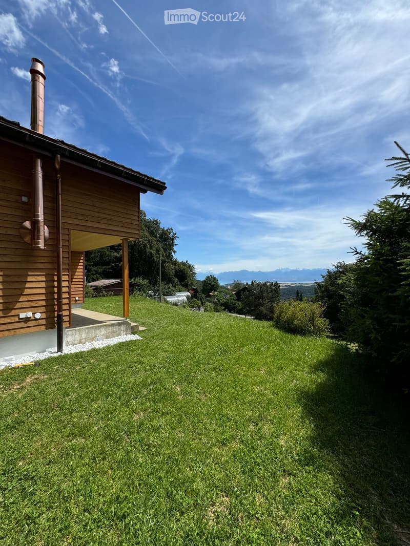 Lush green lawn in front of a wooden house with a raised ground floor, surrounded by trees and a scenic mountain view in the background.