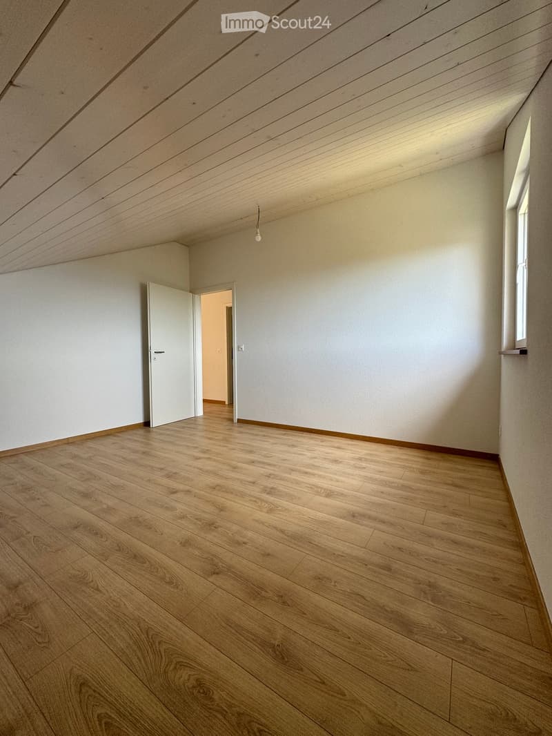 Empty room with wooden flooring, white walls, and a slanted wooden ceiling. Features a single window and an open doorway leading to another room.