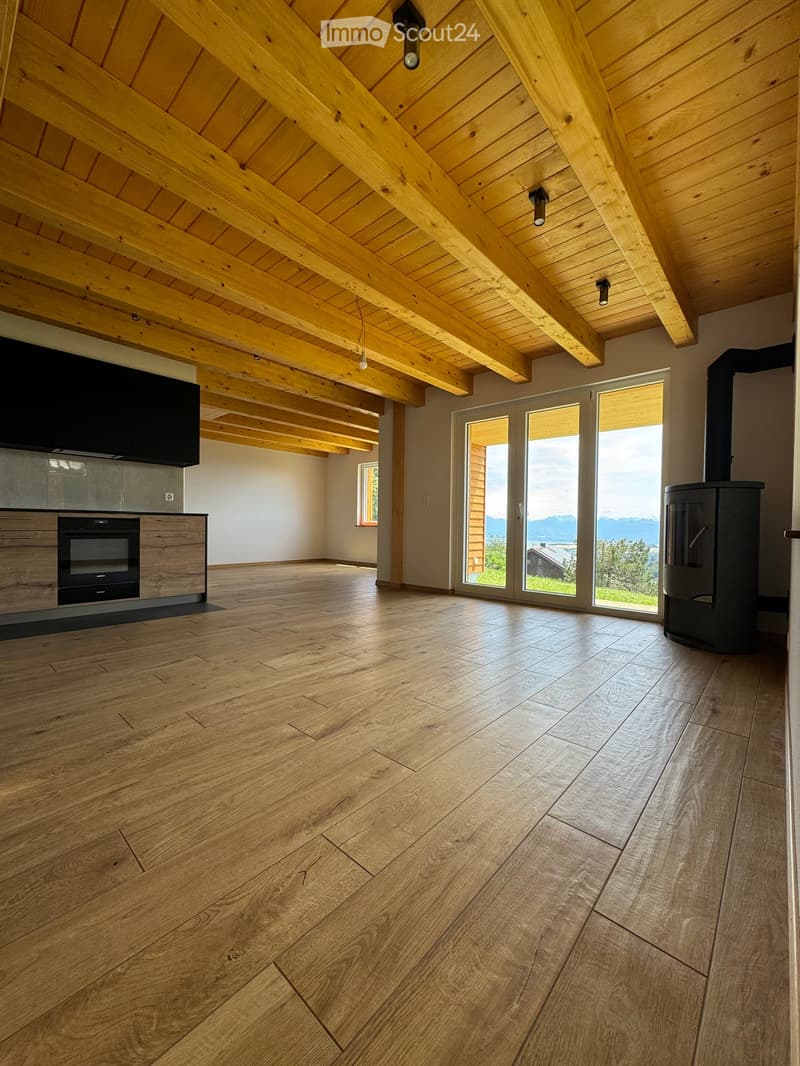 Open-plan living and dining area with exposed wooden beams, hardwood flooring, large windows offering a scenic view, and a modern oven and wood stove.