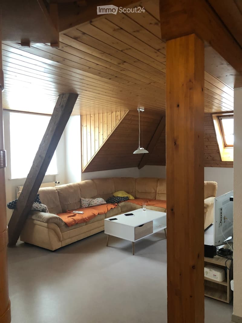 Living room with wooden slanted ceiling, large windows, beige sofa with orange cushions, white coffee table, and TV on a stand.