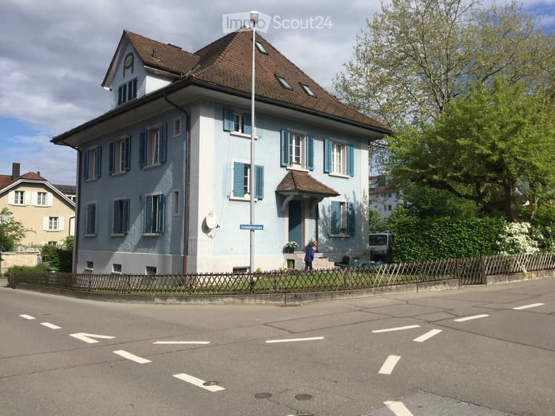 Two-story house with light blue walls, dark blue shutters, and a brown tiled roof. The house features a small porch with a roof and a small garden area in front.