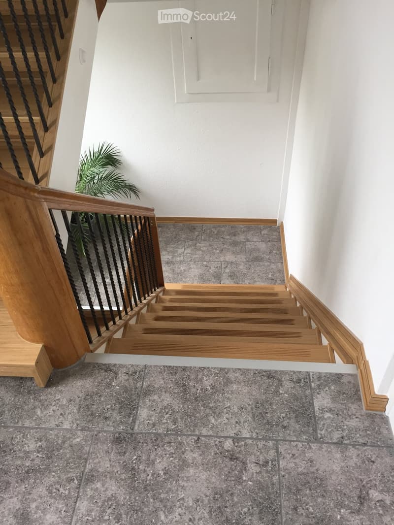Staircase with wooden steps and handrails, grey tiled floor, white walls, and a small green plant in the corner.