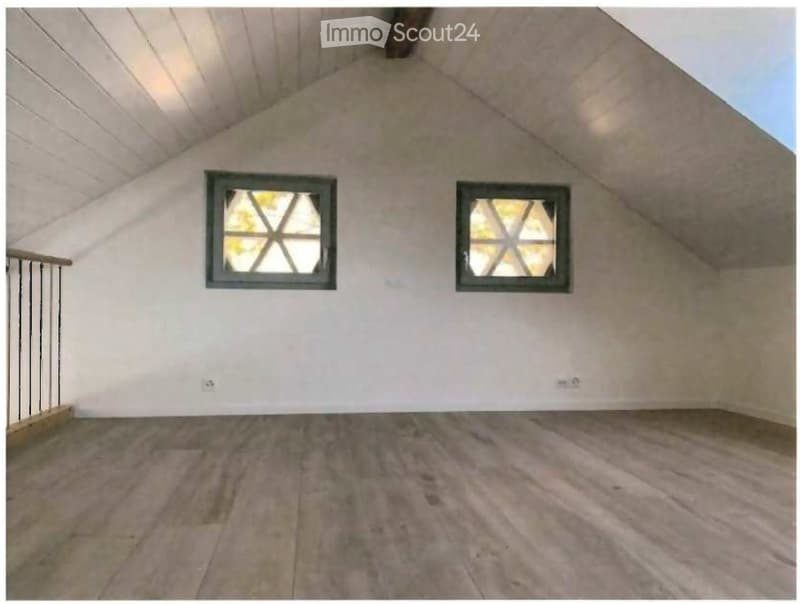 Empty attic space with wooden flooring, white walls, and two windows with decorative glass panes. The ceiling has exposed wooden beams and a sloped design.