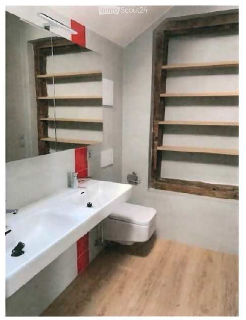 Modern bathroom with a white sink, toilet, and wooden shelves. The walls are light grey with red accents, and the floor is light wood.