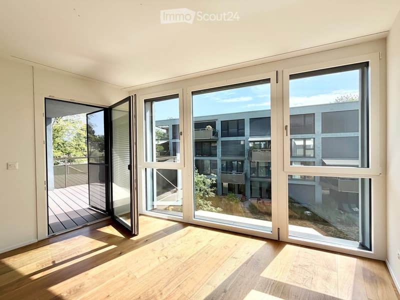 Bright, empty room with wooden flooring and large glass windows and sliding doors that open to a balcony and outdoor area.