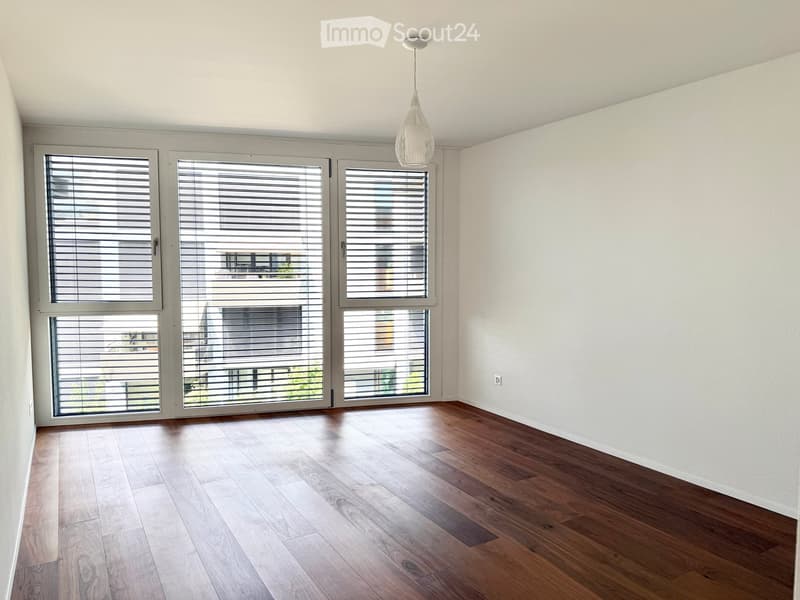 Empty room with wooden flooring, large windows with blinds, and a hanging light fixture.