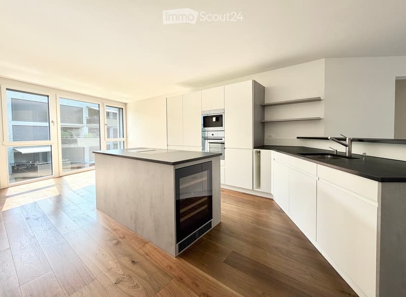 Modern kitchen with white cabinets, a central island, and built-in appliances including a microwave and oven. Features a black countertop, wooden flooring, and large windows with a view of the balcony.