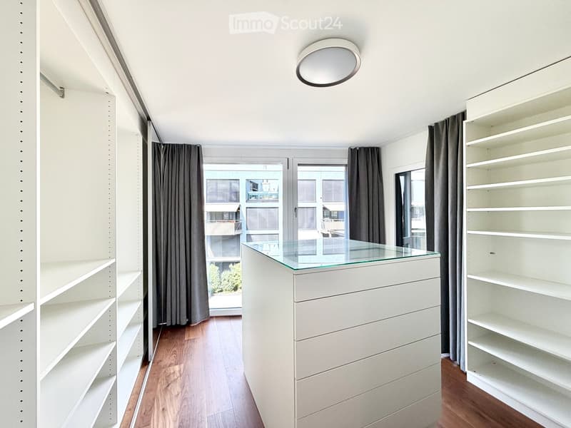 Empty room with white built-in shelving units, a glass-topped cabinet, wooden flooring, and large windows with grey curtains.