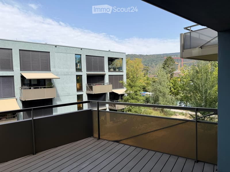 Balcony with wooden decking, glass railing, overlooking a modern building and green landscape.
