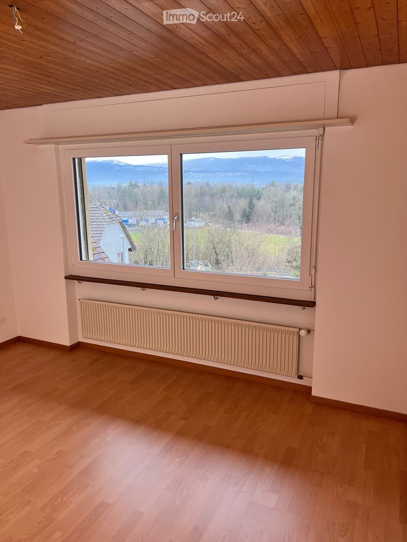 Empty room with wooden flooring, large windows offering a scenic mountain and lake view, radiator, and wooden ceiling beams.
