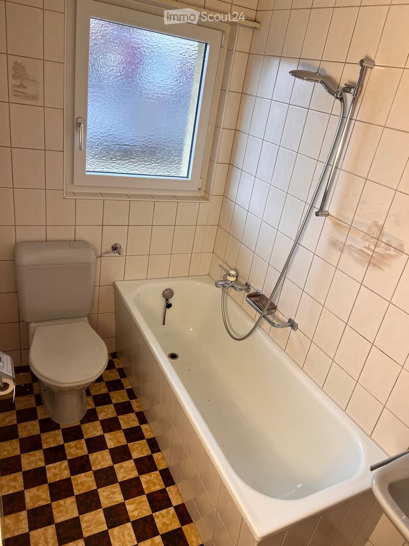 Bathroom with a bathtub, toilet, and showerhead. The walls are tiled, and the floor has a checkered pattern.