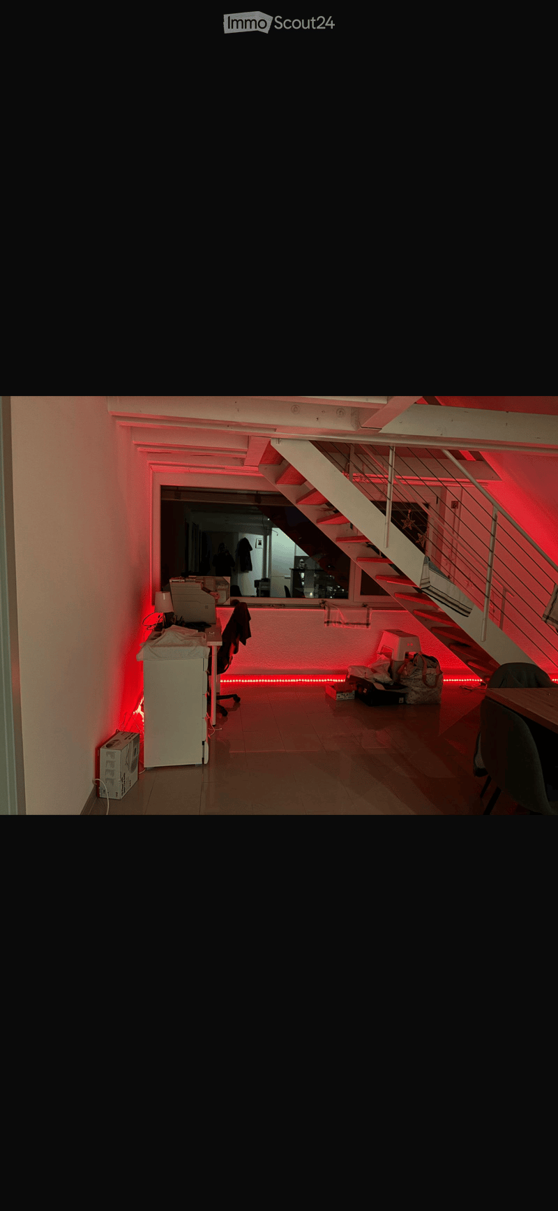 A modern, well-lit room with a staircase leading upwards, a chair, a table, a lamp, and a mirror on the wall.