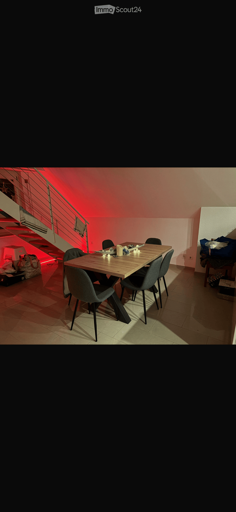 Dining area with a wooden table, four chairs, and a candle on the table. A staircase with red lighting is visible in the background.