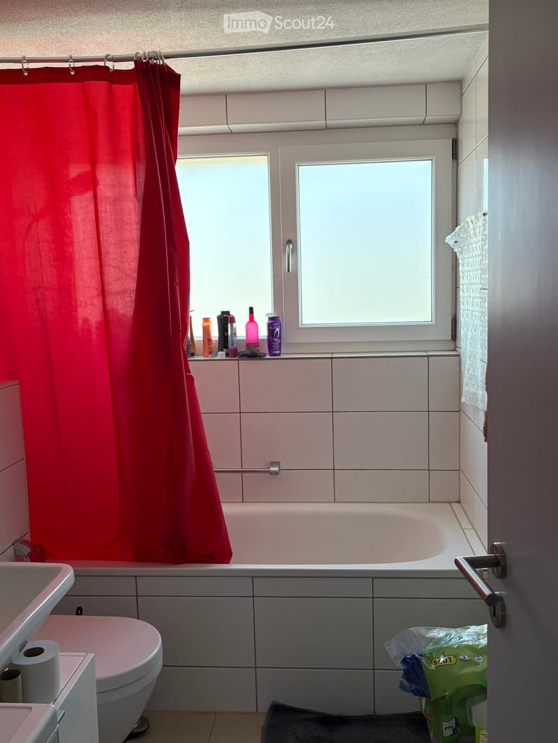 Bathroom with a bathtub, toilet, sink, and shower curtain. Various bathroom products are placed on the windowsill, and a towel hangs on the wall.