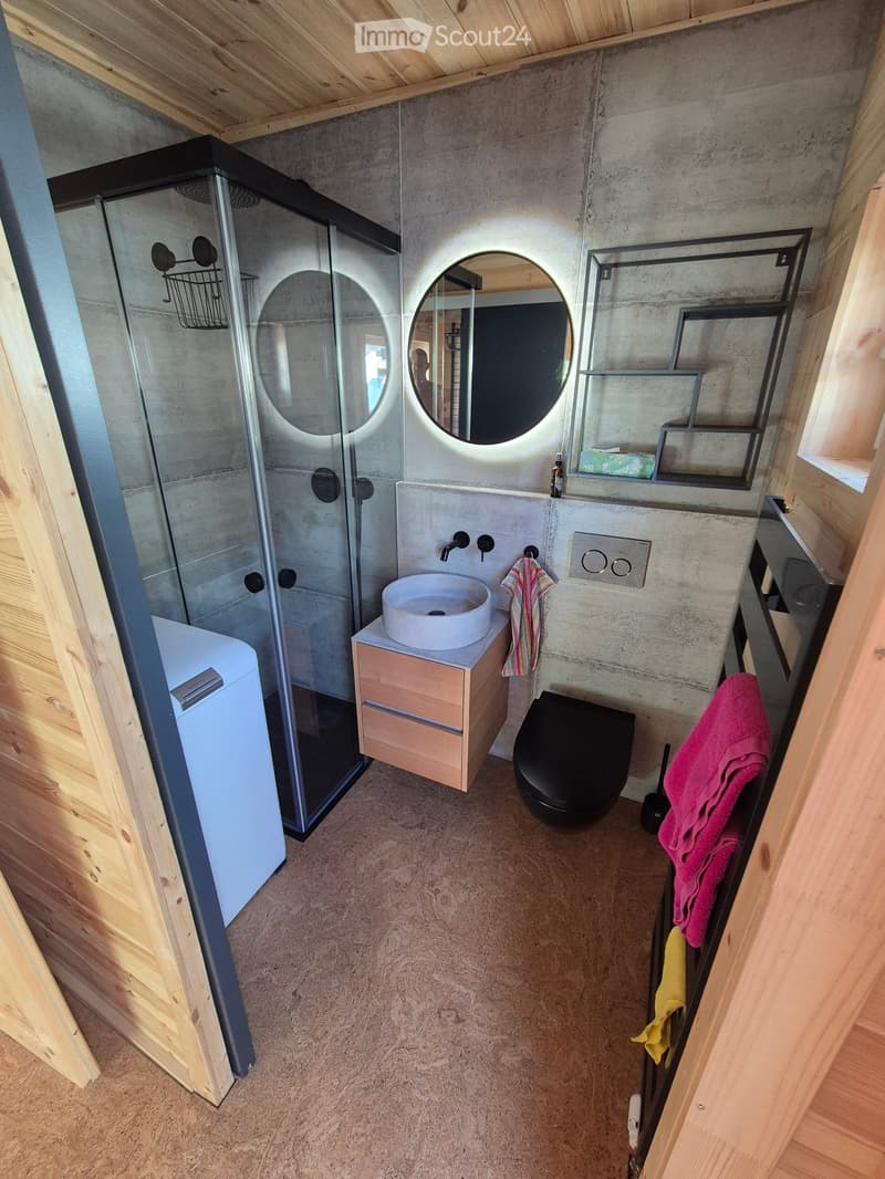 Modern bathroom with a sink, toilet, shower, and mirrors. Features a wooden vanity, a black toilet, and a glass-enclosed shower with a built-in shelf. The room has a concrete wall finish and a wooden floor.