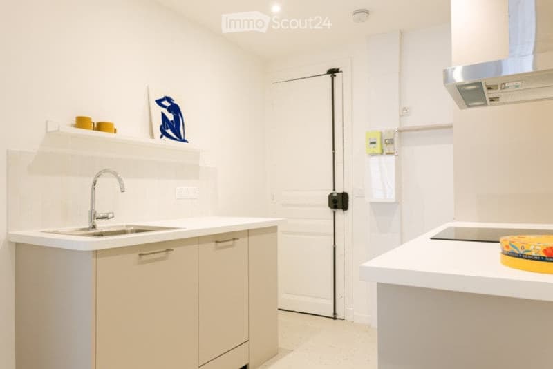 Modern, minimalist kitchen with white cabinets, a stainless steel sink, and a built-in oven. The space features sleek countertops, a small art piece on the wall, and a few decorative items on the shelves.