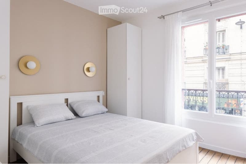Bright bedroom with a white bed, grey pillows, a tall white wardrobe, and two decorative wall-mounted hat racks. The room features wooden flooring and large windows with white curtains, offering a view of a cityscape with balconies.