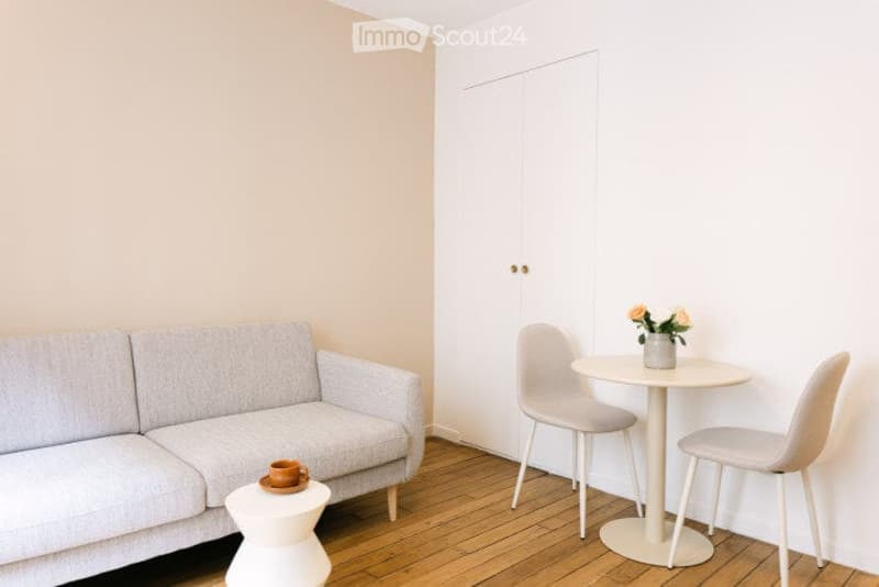 A small, modern living room with a grey sofa, a white round table with two chairs, a small stool, and a vase with flowers on the table.