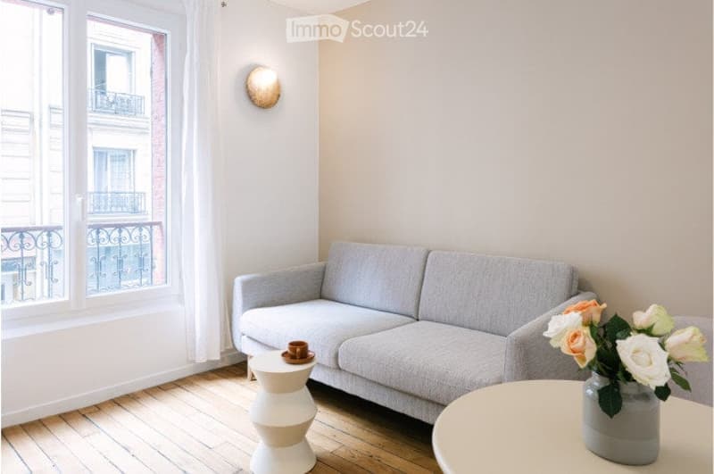 Modern living room with wooden floor, large window with view, grey sofa, two white stools, round table with vase of flowers.