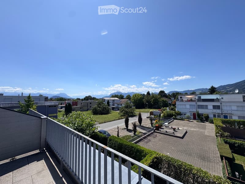 Balcony overlooking a residential area with buildings, trees, and a clear blue sky with distant mountains.