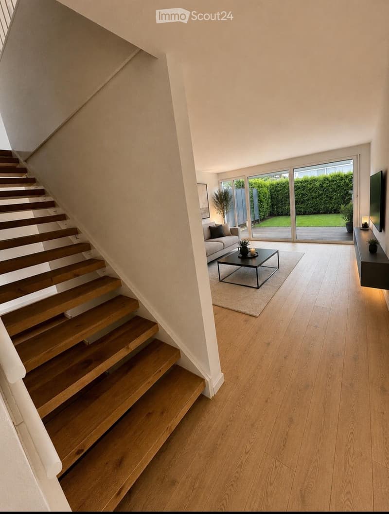 Modern living room with wooden flooring, a staircase leading upwards, a sofa, coffee table, and a TV screen. Large windows provide natural light and a view of the garden.