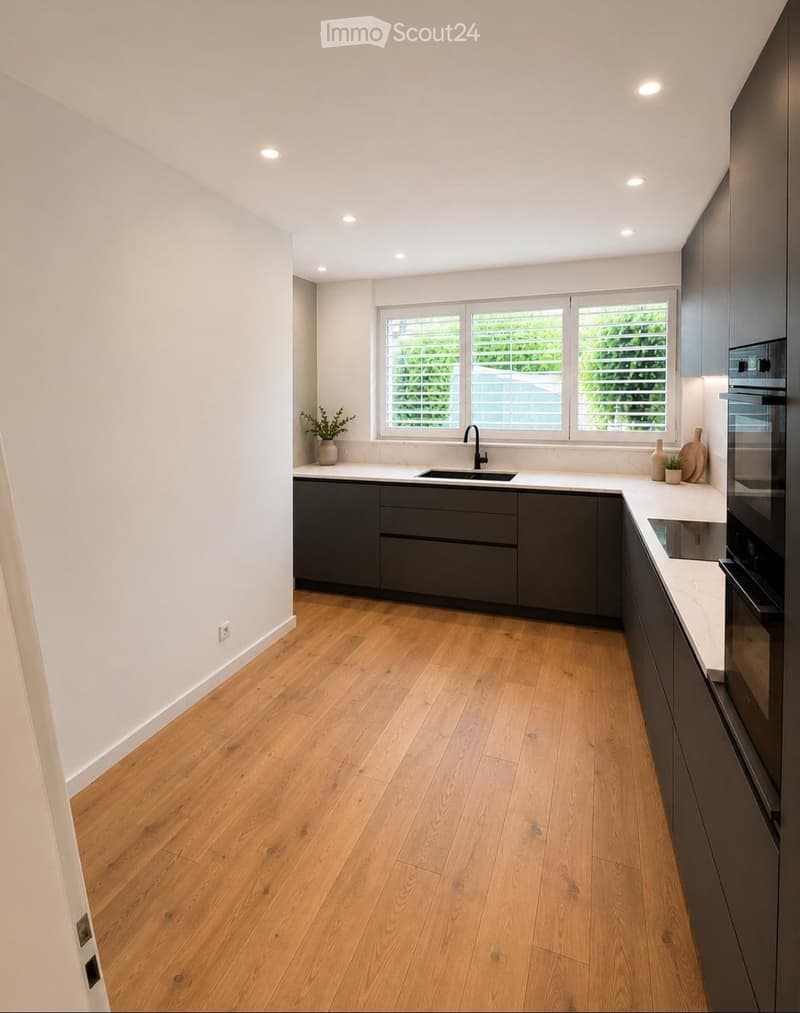 Modern kitchen with light wooden flooring, dark grey cabinets, white countertops, and a central island with a sink and faucet. Features include recessed ceiling lights, a window with blinds, and small decorative plants on the counter.