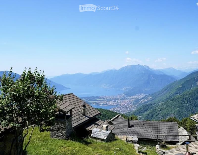 A rustic stone house with a tiled roof, situated on a grassy hillside with a panoramic view of a lake and surrounding mountains.