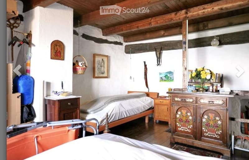 Cozy bedroom with wooden beams, a single bed, nightstand, dresser, mirror, and various decorative items including a wicker basket and picture frames.